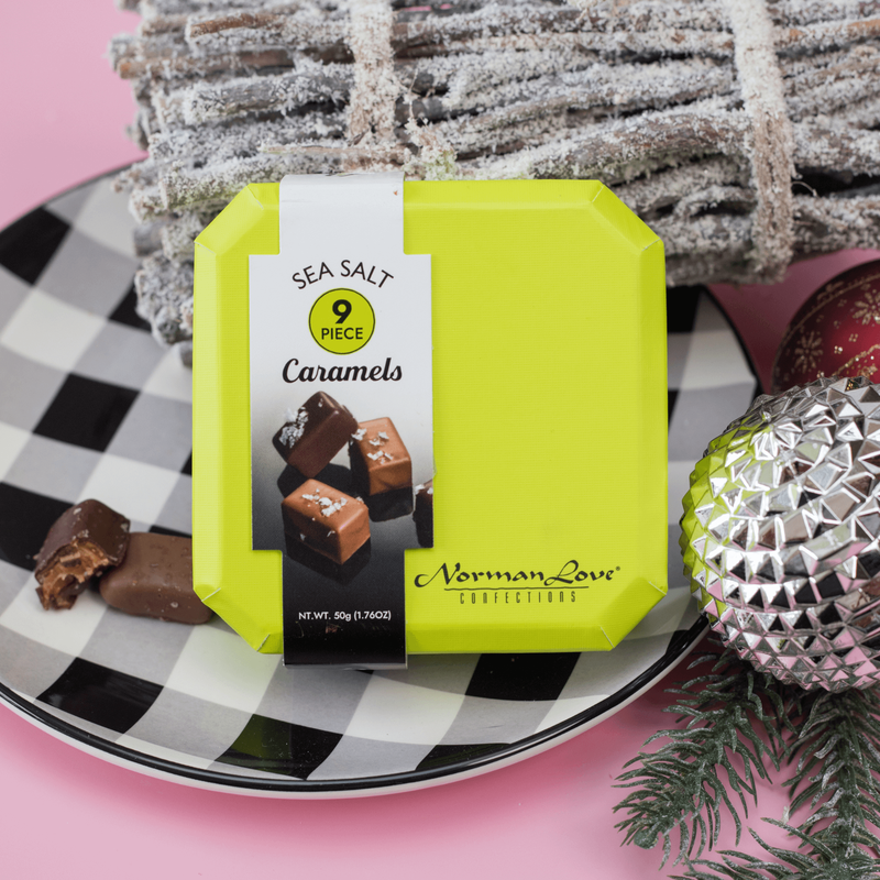 A box of Sea Salt Caramels displayed on a black and white checkered plate. Two caramels, one dark chocolate and one milk chocolate, sit outside the box. A bundle of snow-covered sticks and two ornaments bring forth the spirit of the season. 